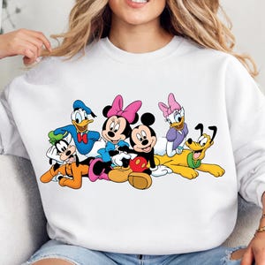 May include: White sweatshirt featuring a colorful graphic of Disney characters, including Mickey and Minnie Mouse, Donald Duck, Goofy, Daisy Duck, and Pluto. The cartoon characters are in a group, with vibrant colors and distinct features.