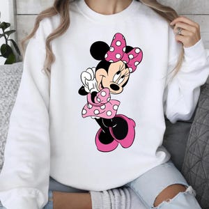 May include: White sweatshirt featuring a cartoon illustration of Minnie Mouse. Minnie Mouse is depicted in a pink polka-dot dress, bow, and shoes. The character is smiling and has black ears. The sweatshirt is a casual style.