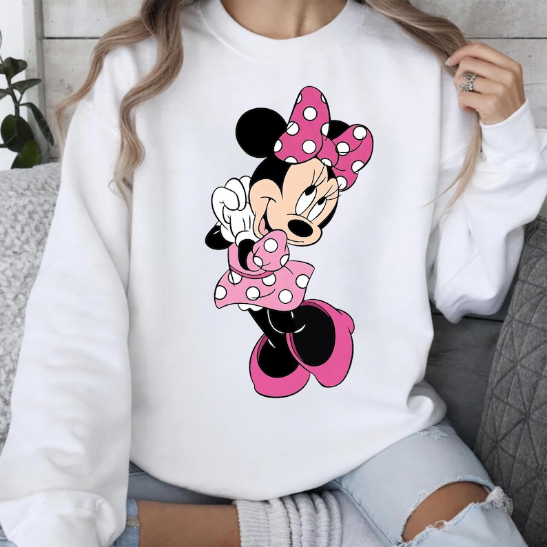 May include: White sweatshirt featuring a cartoon illustration of Minnie Mouse. Minnie Mouse is depicted in a pink polka-dot dress, bow, and shoes. The character is smiling and has black ears. The sweatshirt is a casual style.