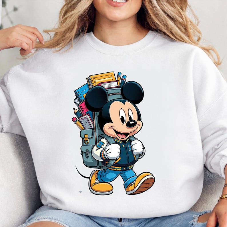 Op de afbeelding: Witte sweatshirt met een cartoon illustratie van Mickey Mouse die een varsity jas, blauwe jeans en gele schoenen draagt, met een rugzak vol schoolspullen. Het ontwerp is kleurrijk en levendig, perfect voor de terugkeer naar school.