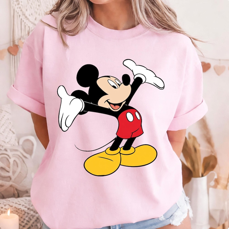 May include: A light pink t-shirt featuring a large graphic of Mickey Mouse. Mickey is depicted with black ears, a black face, white gloves, red shorts, and yellow shoes. The cartoon character is in a celebratory pose.