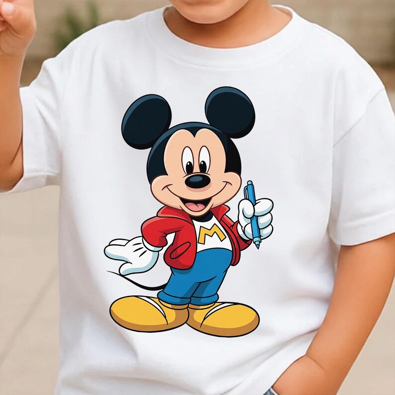 Op de afbeelding: Wit T-shirt met een Mickey Mouse-afbeelding. Mickey draagt een rode jas, een blauwe broek en gele schoenen, en houdt een blauwe pen vast. Het ontwerp bevat de letter "M" op de jas. Een klassiek cartoon-personage-ontwerp.