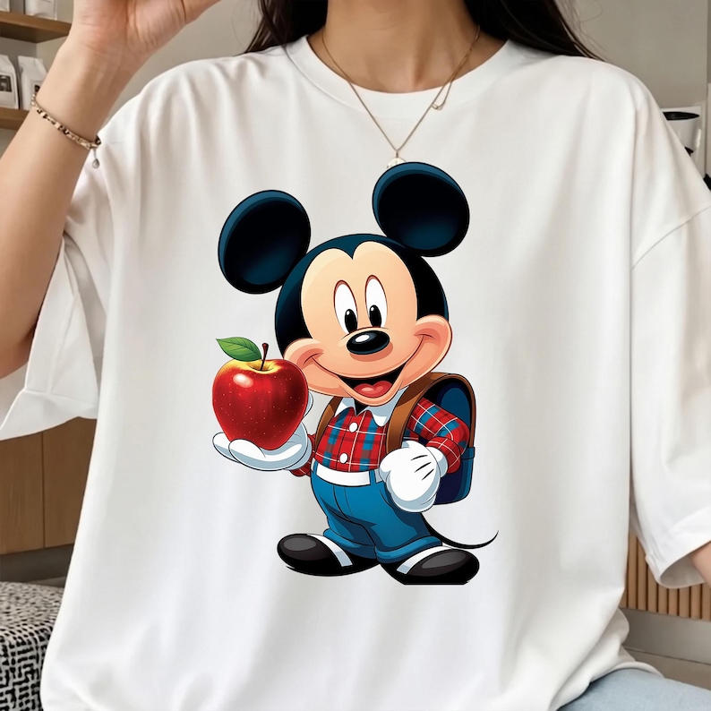 Op de afbeelding: Wit T-shirt met een cartoonafbeelding van Mickey Mouse die een rode appel vasthoudt. Mickey draagt een geruit overhemd, een blauwe broek en een rugzak. Een kleurrijk en levendig ontwerp, perfect voor Disney-fans.