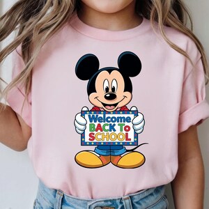 Op de afbeelding: Roze T-shirt met een cartoon Mickey Mouse die een bord vasthoudt met de tekst "Welcome Back To School". Mickey draagt een blauwe broek, een rood shirt en gele schoenen. Het bord heeft een blauwe rand met kleurrijke tekst.