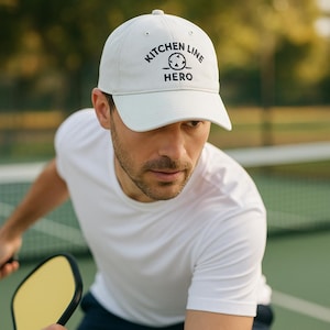 Puede incluir: Gorra de béisbol blanca con el texto negro "KITCHEN LINE HERO". La gorra la lleva una persona que practica deporte en una cancha verde. La persona lleva una camiseta blanca.