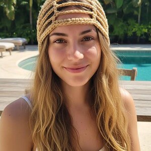 Puede incluir: Una mujer con un sombrero beige de ganchillo con un diseño geométrico. El sombrero está hecho de hilo de color claro. La mujer sonríe y lleva una camiseta blanca. El fondo incluye una piscina y follaje tropical.