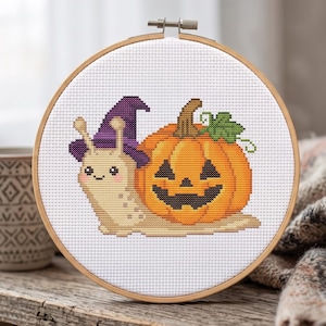May include: A circular wooden embroidery hoop containing a cross-stitch design of a beige snail wearing a purple witch hat next to an orange jack-o'-lantern pumpkin with a green leaf, all stitched on a white fabric background.