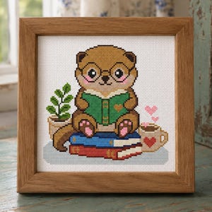 May include: A framed cross-stitch embroidery featuring a brown otter wearing glasses, reading a green book while sitting on a stack of blue and red books. Beside the otter is a small potted green plant and a mug with pink heart-shaped steam.