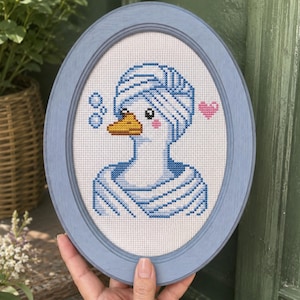 May include: Oval, light blue framed cross-stitch art featuring a duck wearing a blue towel and bubbles. The duck has a yellow beak, pink cheeks, and a pink heart. The frame is a light blue color.