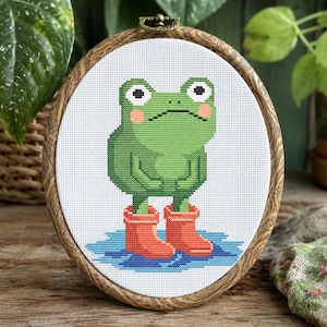 May include: A cross-stitch embroidery piece in a round wooden hoop featuring a green frog with pink cheeks wearing orange rain boots, standing in a blue puddle on a white fabric background. The frog has large white eyes with black pupils.