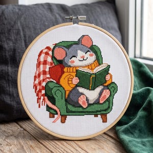 May include: A cross-stitch embroidery in a round wooden hoop featuring a gray and white mouse wearing a yellow sweater, sitting in a green armchair while reading a green book, with a red and white checkered blanket draped over the side of the chair.