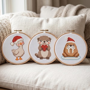 May include: Three circular wooden embroidery hoops containing cross-stitch patterns on white fabric. The left hoop shows a duck in a red Santa hat, the center shows an otter holding a red heart, and the right shows a walrus in a Santa hat.
