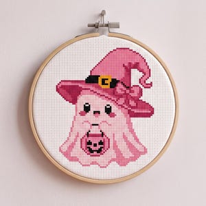May include: A cross-stitch embroidery in a round wooden hoop featuring a light pink ghost wearing a matching pink witch hat with a black band and yellow buckle. The ghost holds a small pink jack-o'-lantern bucket with a black face design.