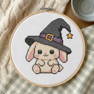 May include: A cross-stitch embroidery in a circular wooden hoop featuring a cream-colored bunny with pink ears and cheeks wearing a dark gray witch hat with a purple band, a yellow buckle, and a small yellow star dangling from the pointed tip.