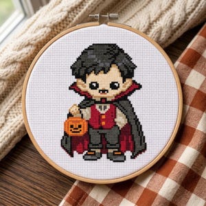 May include: A cross-stitch embroidery in a round wooden hoop featuring a pixelated vampire character. The figure has dark hair, a red vest, and a black cape with a red lining. The vampire holds a small orange jack-o'-lantern bucket on a white fabric.