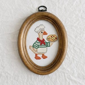 May include: An oval wooden frame containing a cross-stitch embroidery of a goose wearing a white chef hat and a green and white striped shirt with a red neckerchief, holding a round pizza with red and green toppings on a white fabric background.
