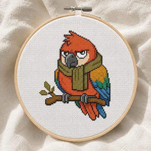May include: A cross-stitch embroidery in a round wooden hoop featuring a colorful parrot with orange, blue, and green feathers. The parrot wears a dark olive green scarf and is perched on a brown branch, depicted with a grumpy facial expression.