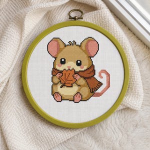 May include: A circular embroidery hoop containing a cross-stitch pattern of a light brown mouse with pink ears and feet. The mouse wears a dark orange scarf and holds a single orange autumn leaf. The design is centered on white fabric.
