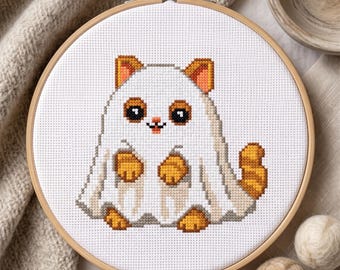 May include: A cross-stitch pattern in a round wooden embroidery hoop featuring a white cat wearing a white ghost costume. The cat has orange ears, orange paws, and an orange tail visible. The design is stitched on white fabric with a grid pattern.