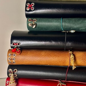 May include: A vertical stack of seven leather-bound journals in various colours including olive green, black, dark green, tan, and red. Each journal features metal eyelets with coloured elastic cords and decorative charms such as hearts, strawberries, and fish.