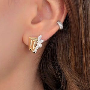 May include: Gold-toned square stud earrings with a leaf-like design of small, clear gemstones. A small, curved ear cuff with multiple small, clear gemstones is also visible. The earrings are set against a background of brown hair.
