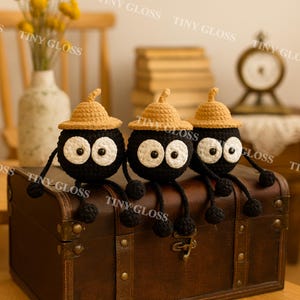 May include: Three handmade crochet figures with black bodies, large white eyes, and tan hats. The figures have black legs and arms and are sitting on a brown leather trunk. The background includes a vase with yellow flowers and a vintage clock.