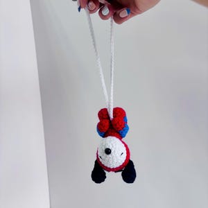 Könnte beinhalten: Ein gehäkelter Hängeschmuck in Form eines weißen Hundes mit schwarzen Ohren, einer schwarzen Nase und geschlossenen Augen, der ein rot-blaues Superhelden-Outfit trägt. Die Figur hängt an einer weißen Häkelschlaufe und ist kopfüber positioniert.