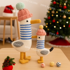 Może przedstawiać: Dwie ręcznie robione figurki mew z szydełka. Większa mewa ma na sobie różową czapkę z zielonym pomponem, a mniejsza żółty pompon. Obie mają niebieskie paski na ciele, żółte dzioby i stopy oraz szare skrzydła.