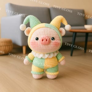 May include: A handmade crochet piglet doll wearing a jester-style outfit. The piglet has a pink snout and cheeks, black eyes, and a two-toned green and yellow hat with white pom-poms. Its outfit is green and yellow, with a white ruffled collar.