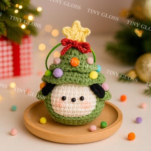May include: A handmade crochet Christmas tree ornament. The green tree is adorned with colorful pom-poms and topped with a yellow star and red bow. The ornament has a white face with black eyes and ears. It sits on a wooden plate.