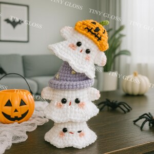 Może przedstawiać: Stos trzech szydełkowych duchów, każdy z innym kapeluszem. Górny duch ma na sobie pomarańczowy kapelusz z dyni, środkowy duch fioletowy kapelusz czarownicy. Czarne oczy i różowe policzki. Dekoracja na Halloween.