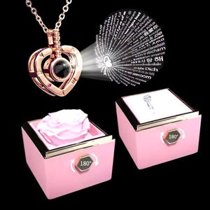 Puede incluir: Un collar con colgante en forma de corazón de oro rosa con función de proyección, acompañado de una caja rosa que contiene una rosa rosa preservada. La caja tiene un borde dorado y un símbolo de rotación de 180 grados. El collar proyecta "Te amo" en varios idiomas.