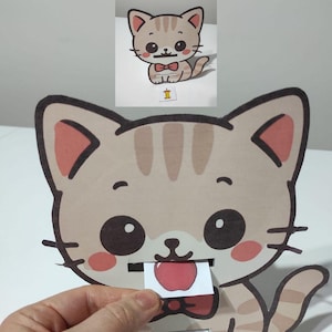 May include: A tan and brown cat-shaped paper craft with a red bow tie and a slot for inserting a small card. The card features a red apple illustration. A smaller version of the cat is visible in the background.