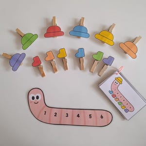 May include: Educational toy set featuring wooden clothespins with colorful hat and boot cutouts. Includes a pink worm-shaped number line from 1 to 6 and a card with a worm illustration. The set is designed for early learning and counting.