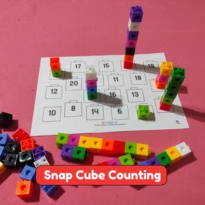 May include: A set of colorful plastic snap cubes and a white activity sheet with numbered squares. The sheet features the text STACK IT and Snap Cube Counting. Several cubes are stacked into vertical towers on the sheet, while others are loose.