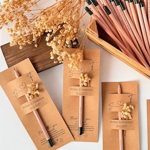 May include: A collection of pencils with green tips in a wooden crate, alongside individual pencils attached to kraft paper cards. The cards are decorated with dried flowers and personalized text, suggesting a wedding favor or event giveaway.