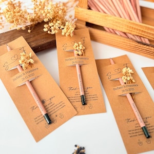 May include: Wedding favors featuring pink pencils with black tips, attached to brown cards with dried flowers. The cards are labeled with names and a date, and the text "Thank you". A wooden box holds more pencils.