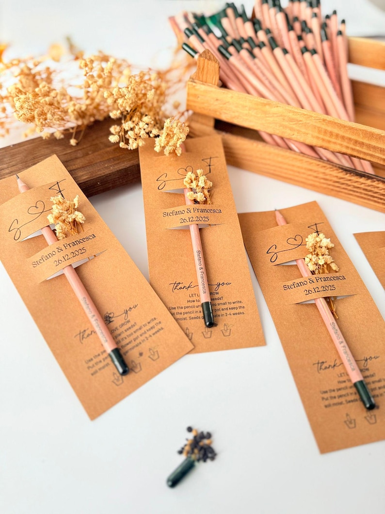 May include: Wedding favors featuring pink pencils with black tips, attached to brown cards with dried flowers. The cards are labeled with names and a date, and the text "Thank you". A wooden box holds more pencils.