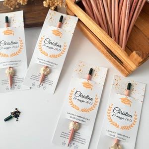 May include: Graduation-themed pencils with attached seed packets. Each pencil is light brown with a black tip and a small seed packet. The pencils are displayed in a wooden box and arranged with cards that say "Christina 20 maggio 2023".