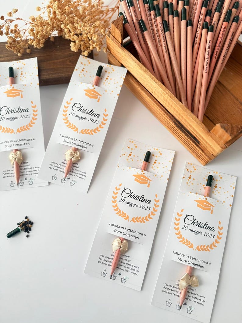 May include: Graduation-themed pencils with attached seed packets. Each pencil is light brown with a black tip and a small seed packet. The pencils are displayed in a wooden box and arranged with cards that say "Christina 20 maggio 2023".