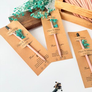 Pu&ograve; includere: Matite rosa con punte nere sono esposte con etichette di carta kraft e piccoli bouquet di fiori blu. Le etichette riportano "Chloe & James 17.10.2025" e "Happily Ever After". Una scatola di legno contiene altre matite.