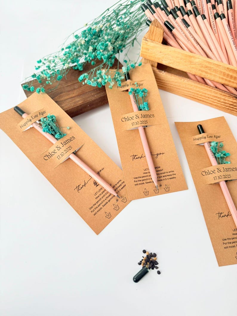 Pu&ograve; includere: Matite rosa con punte nere sono esposte con etichette di carta kraft e piccoli bouquet di fiori blu. Le etichette riportano "Chloe & James 17.10.2025" e "Happily Ever After". Una scatola di legno contiene altre matite.