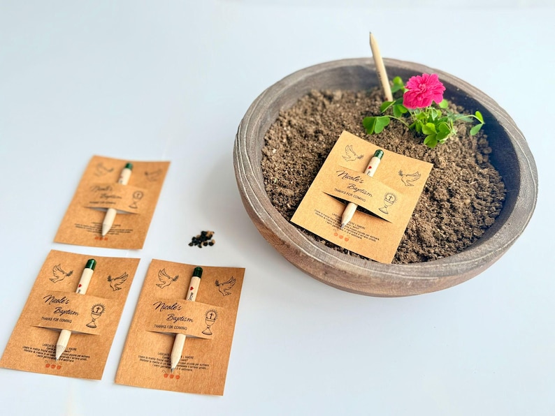 May include: A wooden bowl containing soil, a pink flower, and a small plant. Seed packets with pencils are placed around the bowl. The packets are labelled "Nicole's Baptism" and feature a dove and chalice illustration.