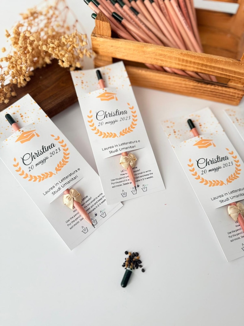 May include: Pencils with seed packets, personalized with the name "Christina" and the date "20 maggio 2023", are arranged on a wooden surface. The pencils are light brown with black tips. The packets have a graduation cap graphic.