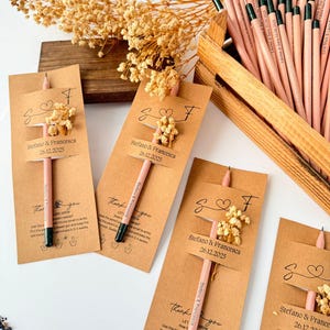 May include: Pencils with attached cards, featuring the names "Stefano & Francesca" and the date "26.12.2025". Each card has a small floral arrangement. The pencils are light pink with black tips. A wooden crate holds additional pencils.