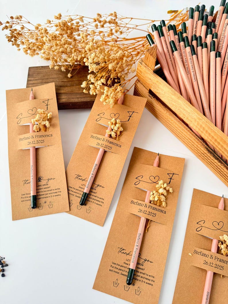 May include: Pencils with attached cards, featuring the names "Stefano & Francesca" and the date "26.12.2025". Each card has a small floral arrangement. The pencils are light pink with black tips. A wooden crate holds additional pencils.