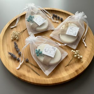 Peut inclure: Trois savons ovales dans des sacs blancs transparents, attachés avec de la ficelle et ornés de petites fleurs bleues et d'étiquettes. Les savons reposent sur un plateau en bois avec de la lavande et des fleurs séchées.