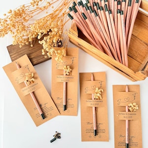 May include: A collection of personalized pencils with the names "Stefano & Francesca" and the date "26.12.2025" printed on them. The pencils are displayed with small floral arrangements and kraft paper tags, suggesting a wedding or event favor.