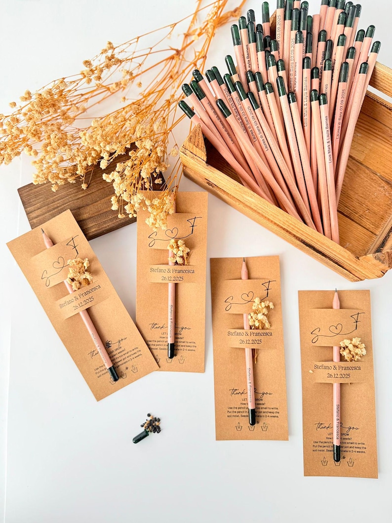 May include: A collection of personalized pencils with the names "Stefano & Francesca" and the date "26.12.2025" printed on them. The pencils are displayed with small floral arrangements and kraft paper tags, suggesting a wedding or event favor.