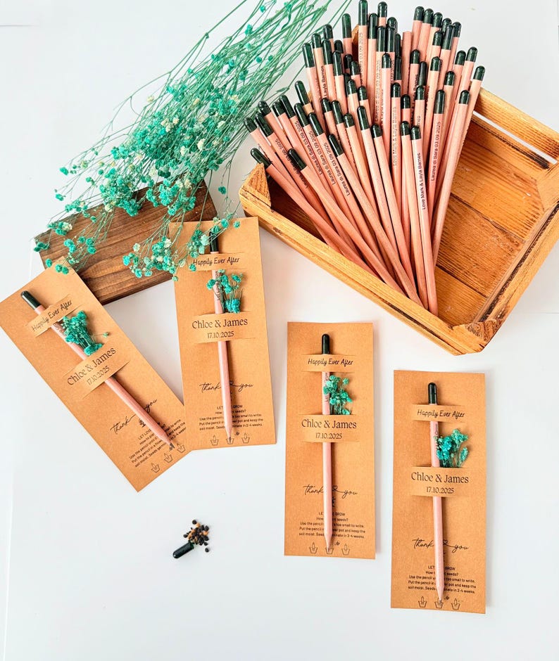 May include: A collection of personalized pencils with the text "Chloe & James 17.10.2025" and "Happily Ever After" printed on them. Each pencil is attached to a brown card with dried blue flowers. A wooden tray holds more pencils.